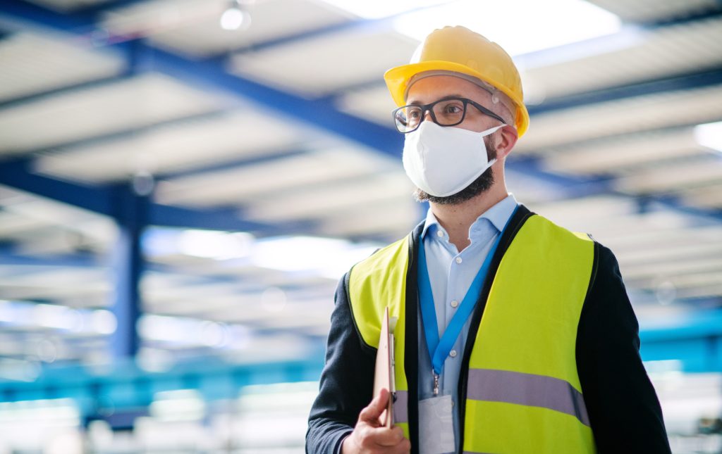 Technician Or Engineer With Protective Mask And Helmet Standing In Industrial Factory.