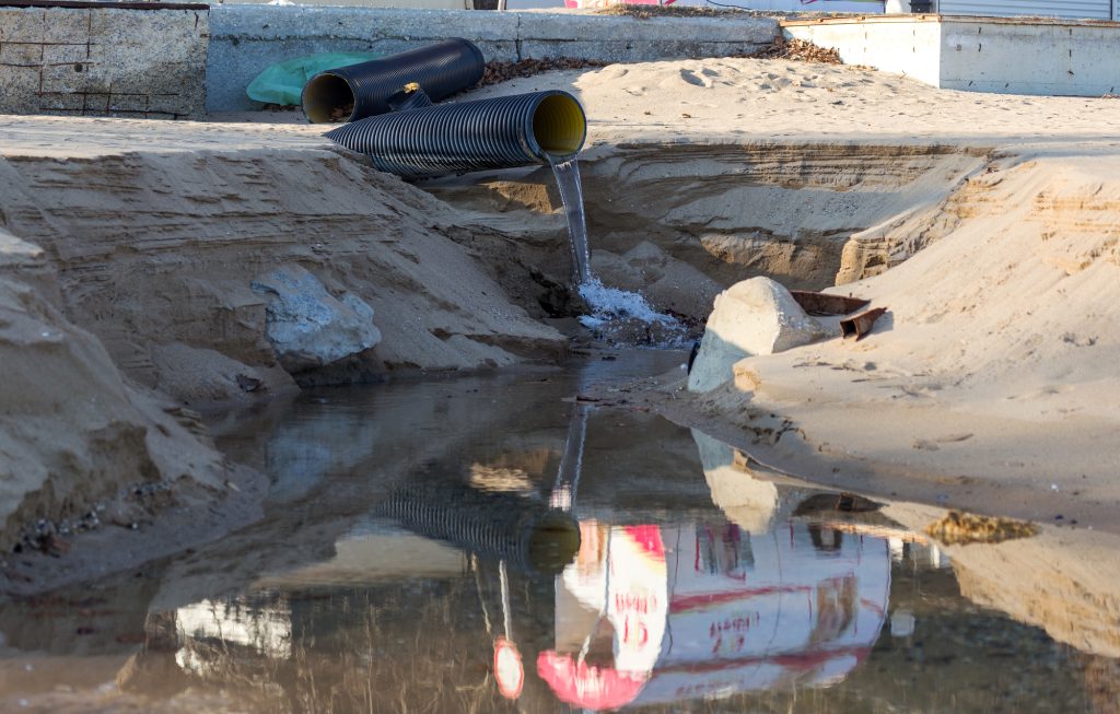 Industrial Wastewater, The Pipeline Discharges Liquid Industrial Waste Into The Sea On A City Beach. Dirty Sewage Flows From A Plastic Sewer Pipe Onto The Sand Of A Sea City Beach